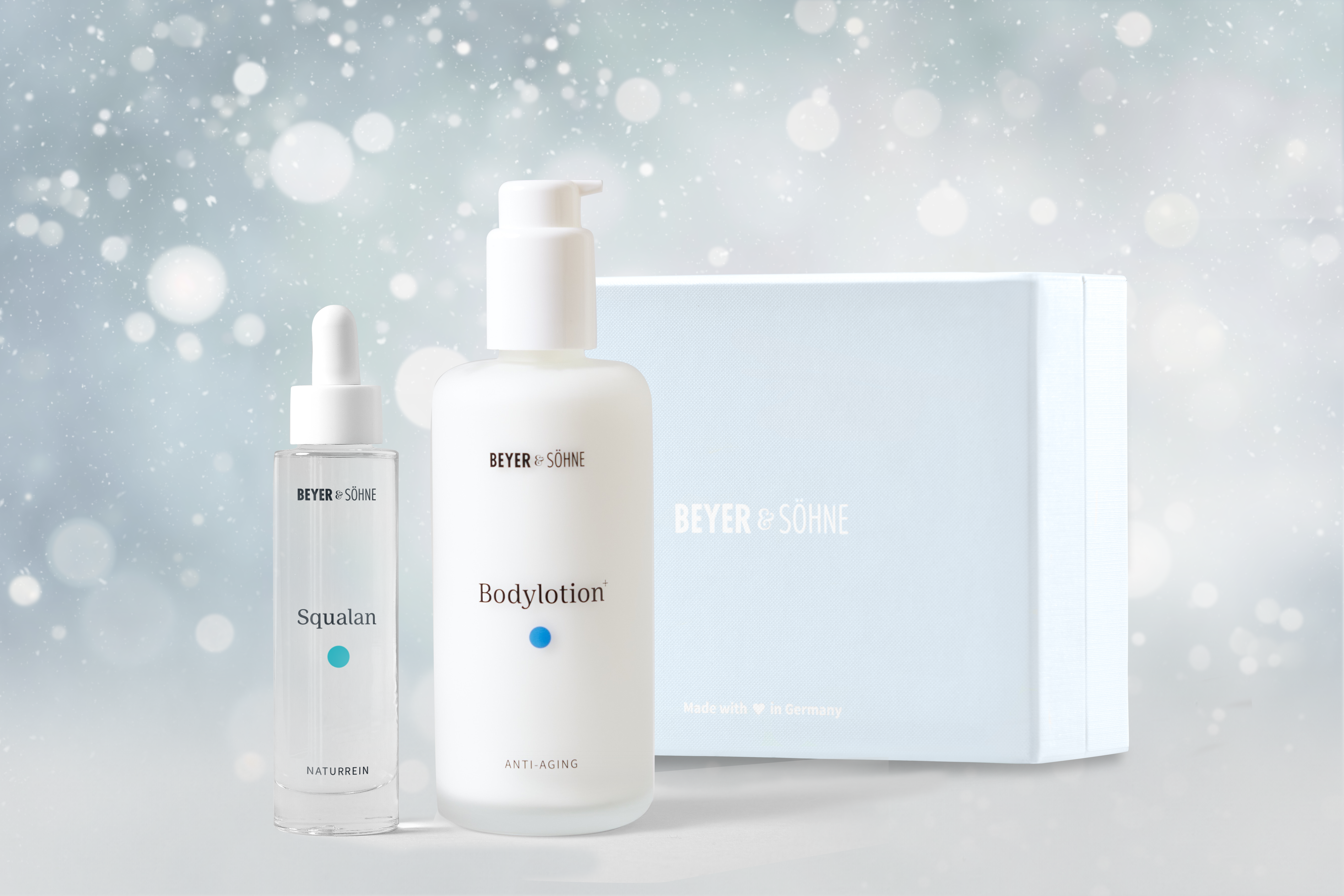 Bodylotion und Squalan in hochwertigen Glasspendern von Beyer & Söhne vor himmelblauer Geschenkbox und weihnachtlich gestimmten Hintergrund mit Schneeflocken.